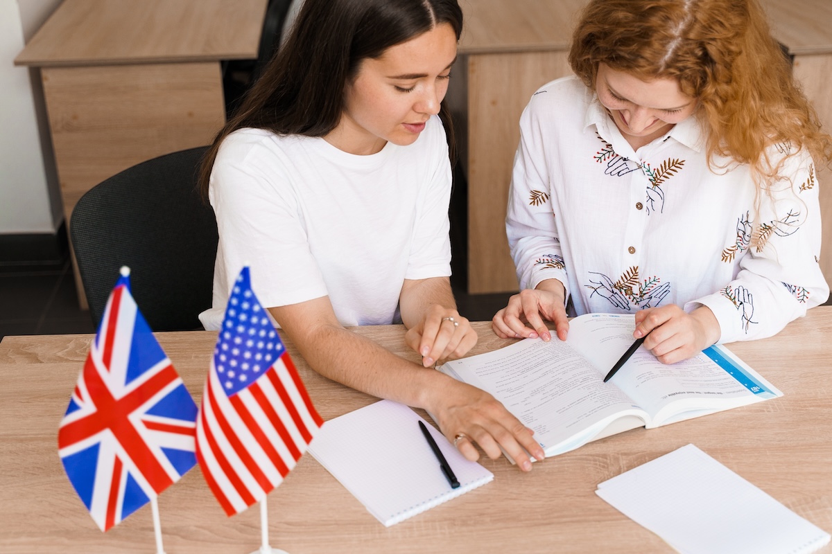 Zwei Teenager beim Englischlernen mit Sprachlehrbuch – Online-Kurs oder traditioneller Englischkurs: Verbessern Sie das Englisch ihrer Kids! Zwei Mädchen mit einem Englischbuch am Schreibtisch, auf dem die US und UK-Flagge stehen – Young-Learners: Englischkurse für Teens.